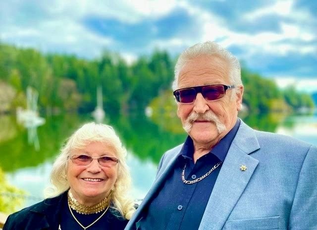 Elderly couple smiling outdoors by a lake with trees in the background.
