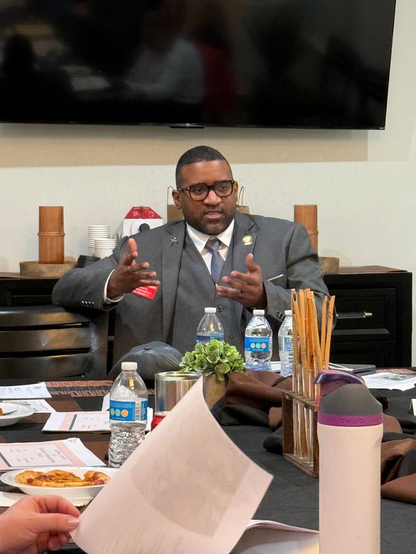 Man in suit and glasses speaking during a meeting at a table with papers and water bottles.