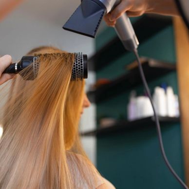 A person blow-drying long blonde hair with a round brush at a salon.