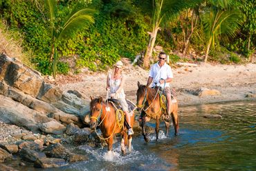 A couple rides horses through shallow water on a tropical beach.