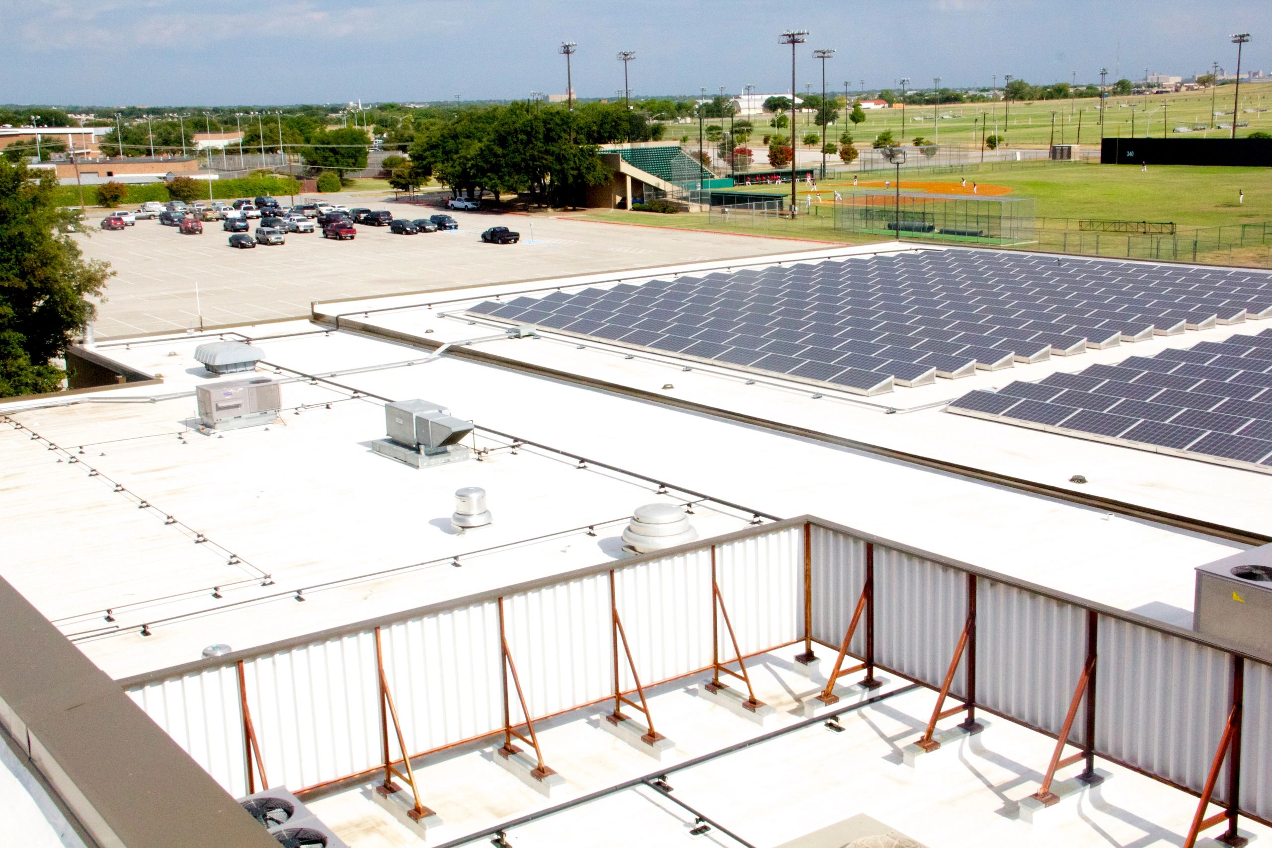 Solar panels on a rooftop overlooking a parking lot and baseball field.
