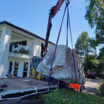 A large rock being lifted by a crane truck near a modern house.