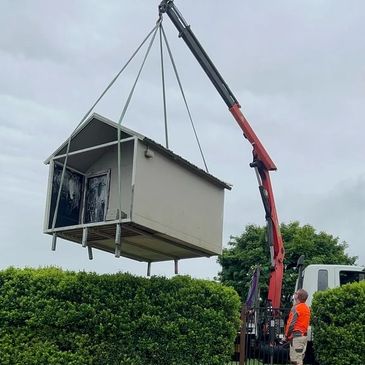 Crane lifting a small house over green bushes with a worker in orange observing.