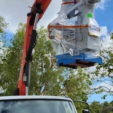 Crane lifting a large machine wrapped in protective plastic outdoors.