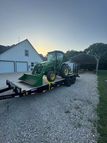 Green John Deere tractor secured on a black trailer outside a white garage at sunset.
