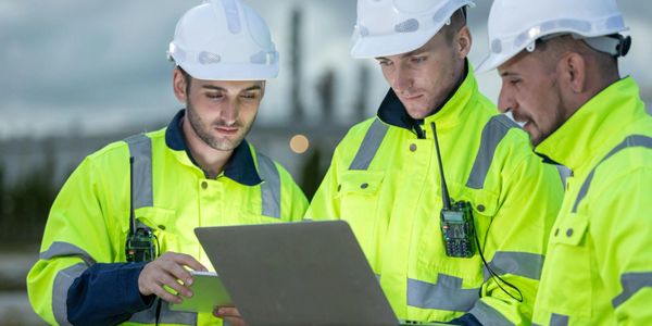 Three construction workers in safety gear using a laptop outdoors.