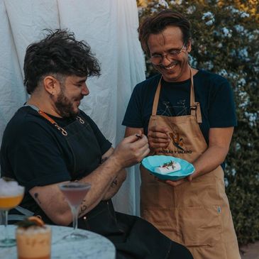 Two men enjoy preparing and tasting food outdoors with happy expressions.