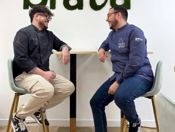 Two chefs in conversation, seated on stools at a table, smiling and relaxed.