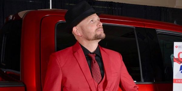 Man in red suit and black hat posing next to a red vehicle.