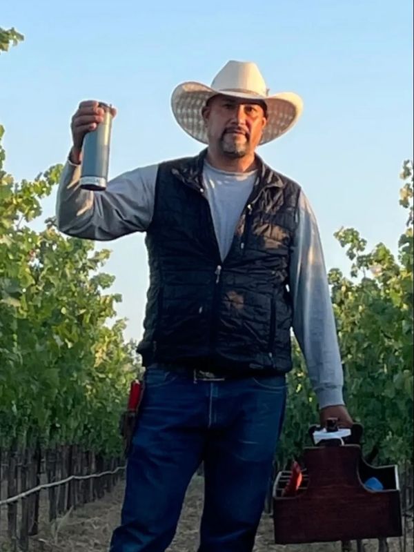 Man in a cowboy hat holding a tumbler and a wooden basket in a vineyard.