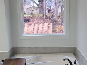 Bathtub with a large window overlooking a backyard with trees and a fence.