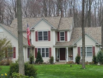 A large beige house with newly painted red shutters, an American flag, and a green yard with trees.