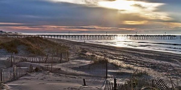 The Sun breaks through the clouds over a pier.  Sand fences  and sea oats are in the foreground.
