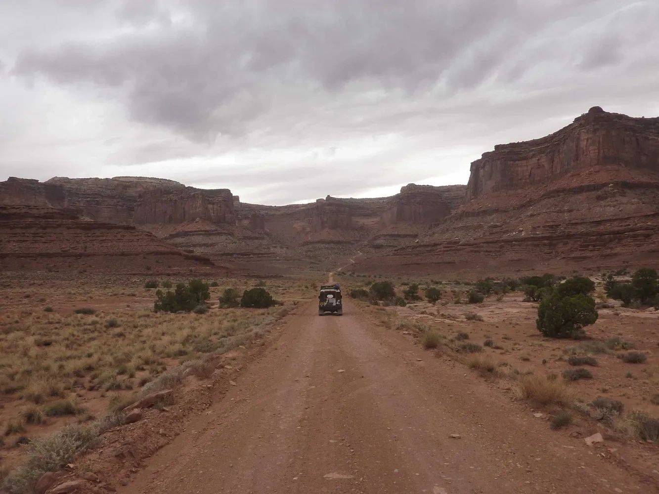 A vehicle driving down a dirt road through a rocky canyon under cloudy skies.