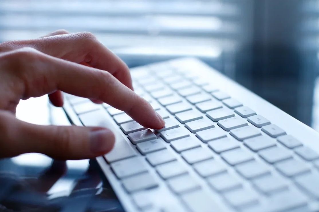 Person's hand typing on a white keyboard. On a glass worktop with blinds in the background. 