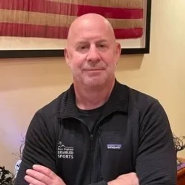 A bald man in a black jacket posing with arms crossed indoors.