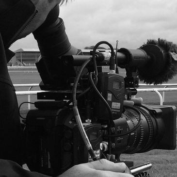 A cameraman filming outdoors with a professional video camera at a racecourse.