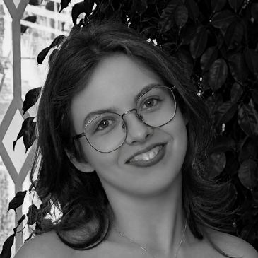 Young woman with glasses smiling in front of leafy background.