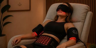 Woman relaxing in a chair wearing a black eye mask and LED therapy devices on arms and abdomen.