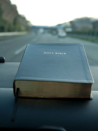 A Holy Bible resting on a car dashboard with a blurred road in the background.