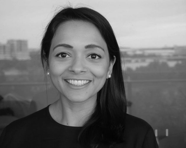 Smiling woman with pearl earrings and dark hair, standing in front of a window with a cityscape background.