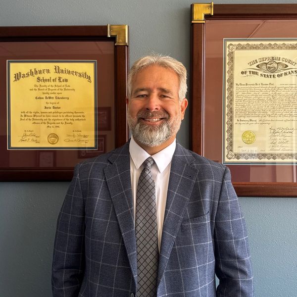 A smiling man in a suit standing between two framed certificates on a wall.