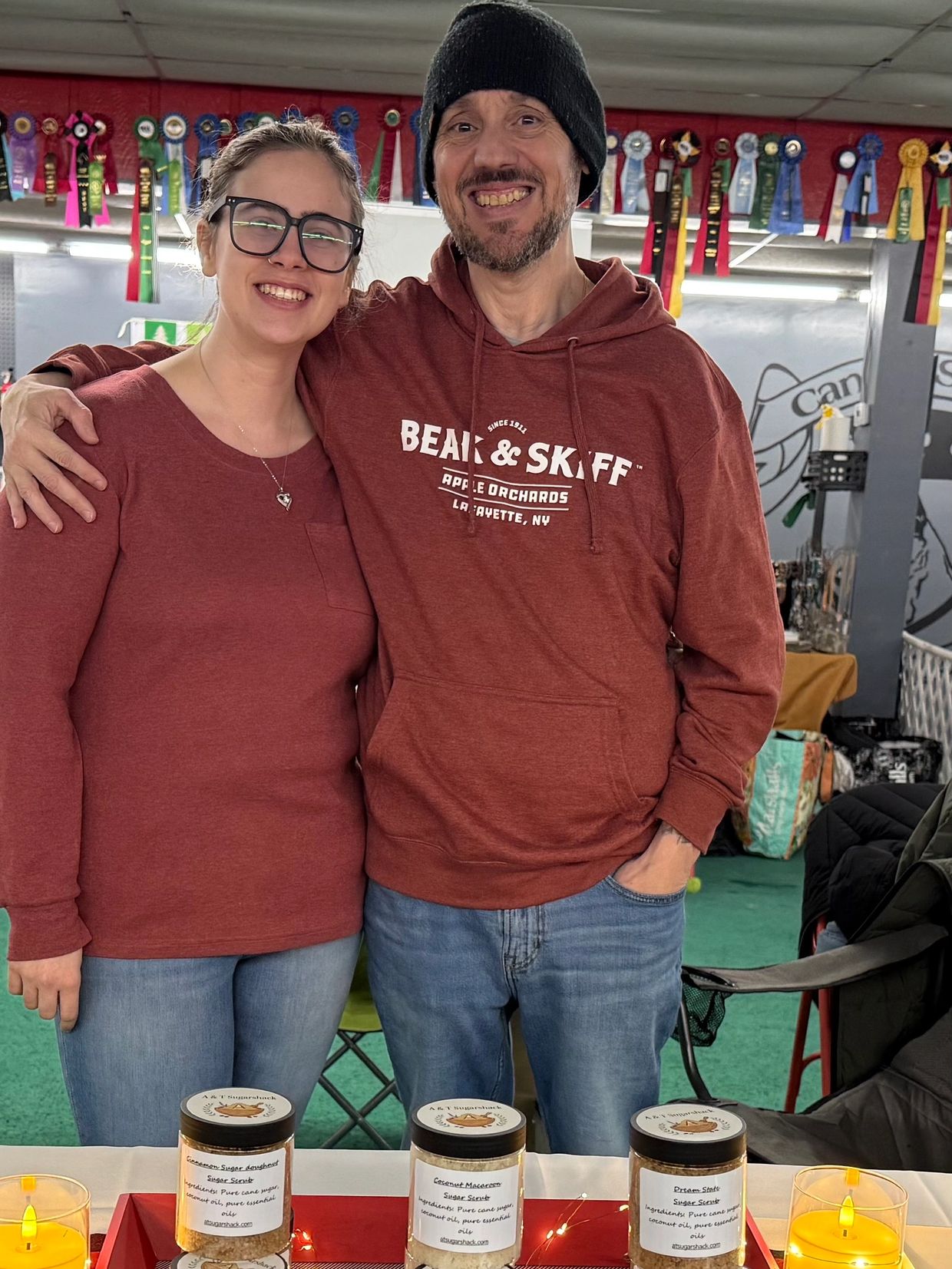 Two smiling people in matching red tops stand behind a table with jars and candles.