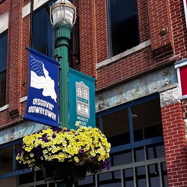 Street lamp with colorful flowers and downtown banners on a brick building.