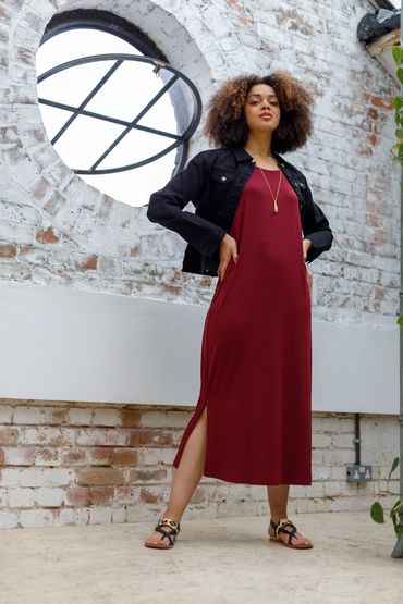 Confident woman in a burgundy dress and black jacket posing indoors by a circular window.