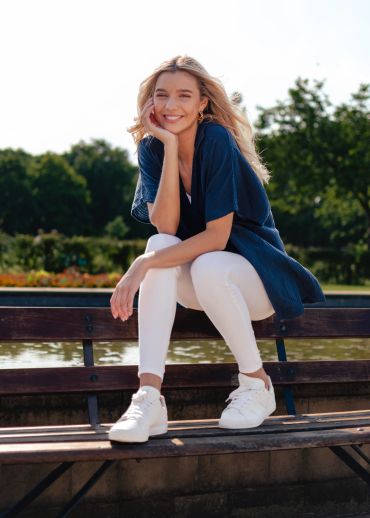Young woman in white pants and navy top smiling on a bench near water.