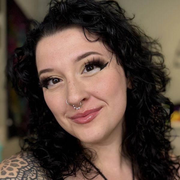 A headshot of a young white woman with black curly hair and intense eye makeup