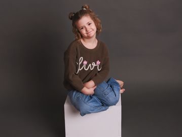 Smiling girl in brown sweater and jeans sitting cross-legged on white box.