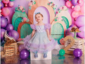 A little girl in a purple dress smiles amidst colorful balloons and decorations.