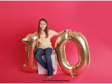 Girl holding gold balloons shaped as 1 and 0 on pink background.