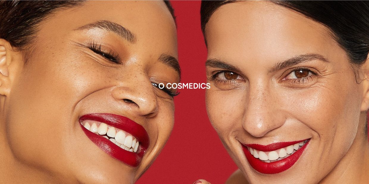 Two women with radiant skin and bold red lipstick smile against a red background.