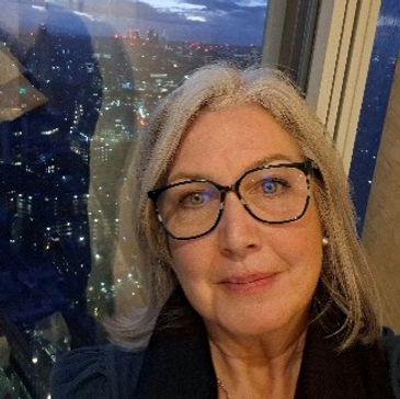 A woman with glasses taking a selfie at night with city lights in the background.