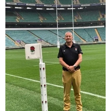 Man standing on a rugby field near a goal post with a rose emblem.