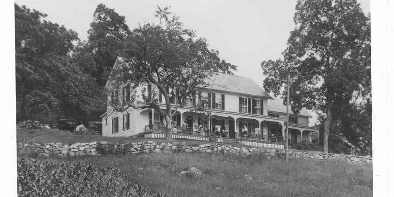 Black and white photo of a large two-story house with a wraparound porch and stone wall.