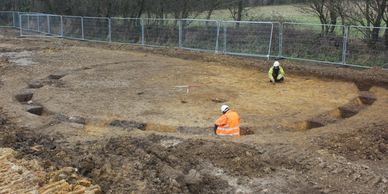 Two workers excavating a large circular archaeological site outdoors.