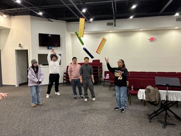 A group of five young people indoors, tossing colorful rectangular objects in the air.