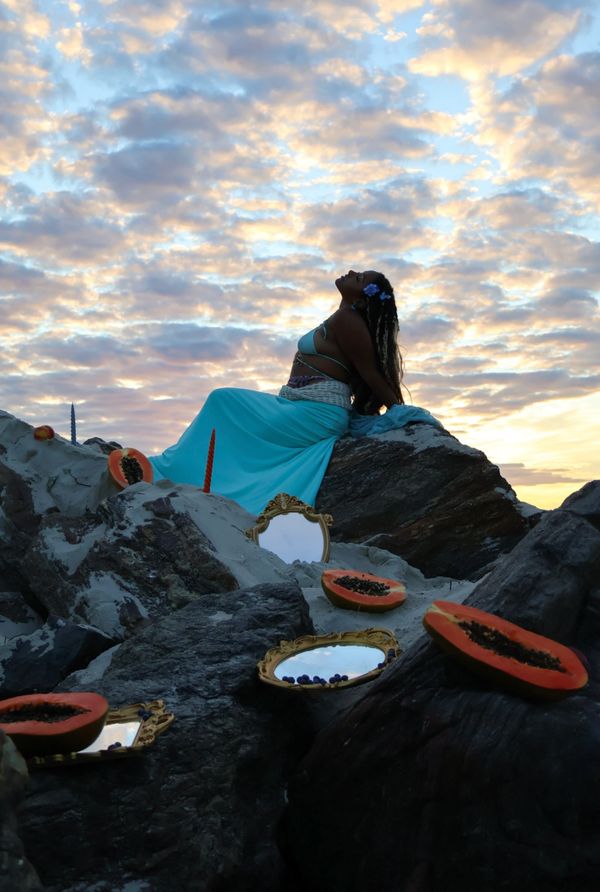 A woman in a blue dress poses on rocks during a cloudy sunset with papayas and mirrors around.