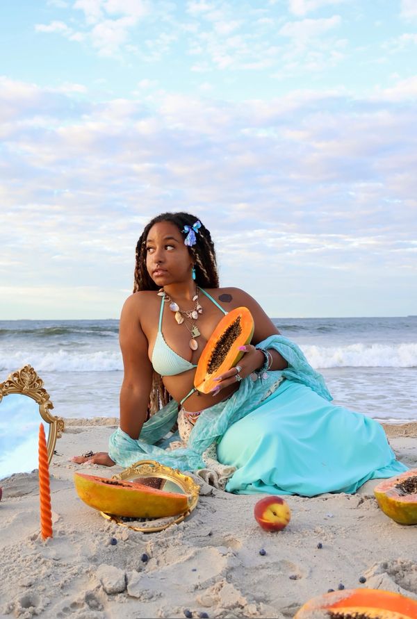 Woman in turquoise attire holds papaya on the beach with seashell necklace and mirror.