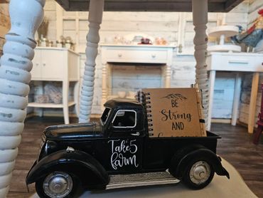 A vintage black toy truck with the words 'Take 5 farm' carrying a notebook and pen inside a rustic room.