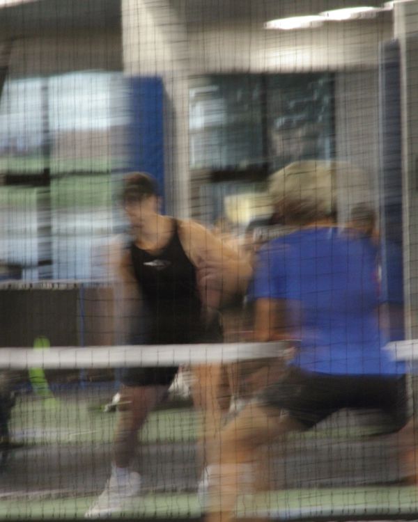 Blurry image of two people playing pickleball indoors behind a net.