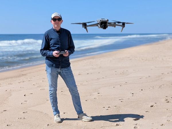 Man flying a drone on a sunny beach with waves in the background.
