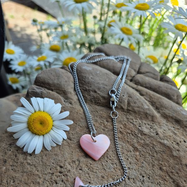 Silver necklace with pink heart pendant near daisies on a rock.