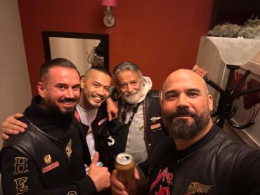 Group of four smiling men in biker attire posing for a selfie indoors.