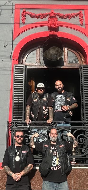 Four tattooed men wearing black leather vests pose on a balcony.