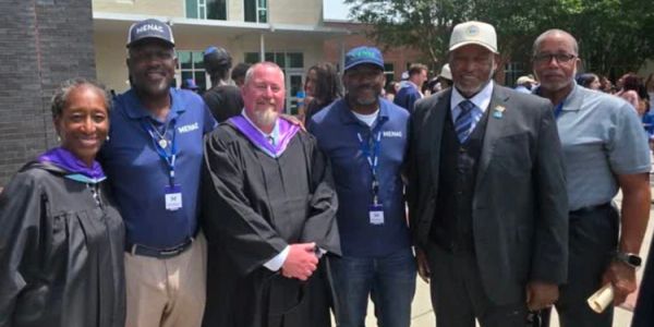 Group of six adults posing outdoors, some in graduation attire.