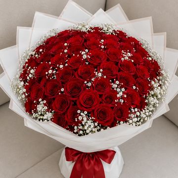 A large bouquet of red roses with white baby's breath, wrapped in white paper and tied with a red ribbon.